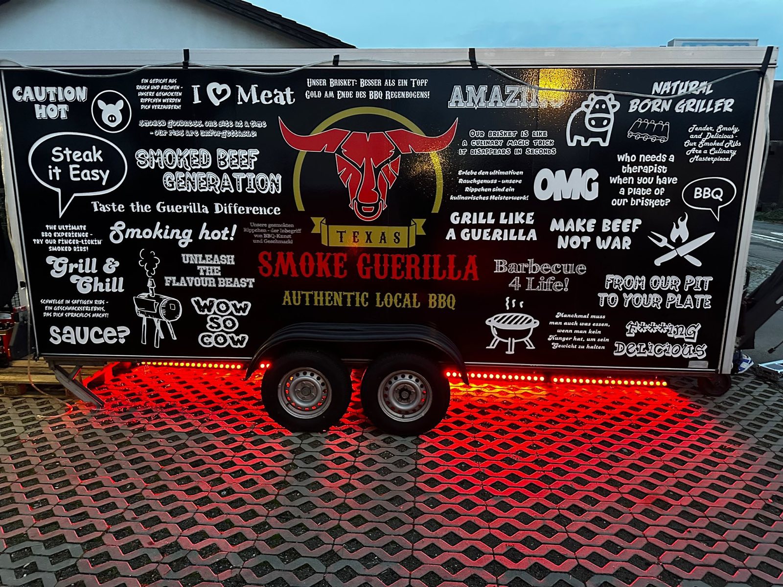 Der schwarze Foodtrailer von Texas Smoke Guerilla mit roter Beleuchtung und BBQ-Sprüchen